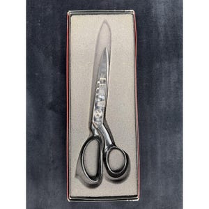 May include: A pair of silver-toned metal scissors with two asymmetrical finger loops, resting inside a rectangular box lined with grey foam. The scissors feature a polished finish and a central pivot screw, presented in a protective storage case.