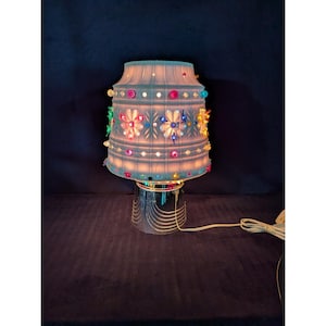 May include: A table lamp featuring a white, textured, bell-shaped glass shade adorned with colorful, multi-shaped glass accents. The shade is illuminated from within, casting a soft glow. The base is a polished, reflective silver-toned metal cylinder.