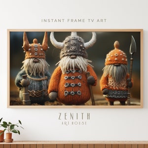Puede incluir: Tres gnomos de punto vestidos como vikingos, con cascos con cuernos y suéteres naranja y grises. Los gnomos están de pie en un entorno de bosque. La imagen está enmarcada con un marco de madera.