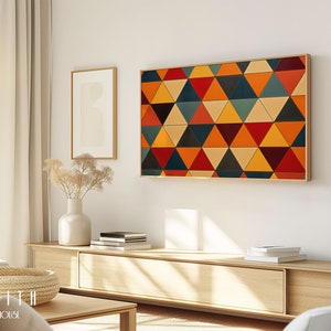 May include: A modern living room interior with a large abstract geometric wall art featuring a pattern of colorful triangles in shades of red, orange, yellow, blue, and green. The art is framed in a natural wood frame and hangs above a light wood entertainment console with a vase of dried flowers and books.