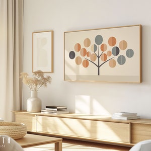 May include: A living room interior with a large framed print of a stylized tree with orange, brown, and blue circles on a cream background. The print is hanging on a white wall above a light wood entertainment center with a vase of dried flowers and books on top.