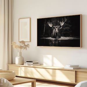 Op de afbeelding: Een zwart-witfoto van een eland die in een waterpartij staat. De eland heeft grote geweien en kijkt recht in de camera. De foto is ingelijst in een dun, gouden lijst en hangt aan een witte muur.