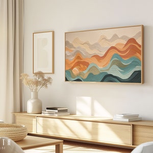 May include: Abstract wall art featuring a layered design of wavy lines in shades of orange, brown, teal, and gray. The artwork is framed in a light wood frame and is hanging on a white wall above a wooden shelf.