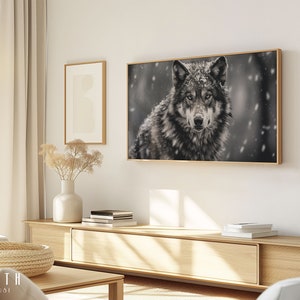 Könnte beinhalten: Ein Schwarzweißfoto eines Wolfes in einem schneebedeckten Wald. Der Wolf blickt direkt in die Kamera mit einem ernsten Ausdruck. Das Foto ist in einem einfachen Holzrahmen gerahmt und hängt an einer weißen Wand.