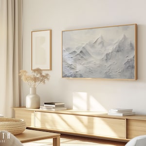 May include: A living room scene featuring a large framed mountain landscape painting with a gold frame. The artwork is predominantly white and grey. Other framed art pieces and a wooden cabinet are also visible.