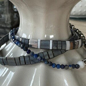 May include: Three bracelets are displayed. One is a dark gray, rectangular bead bracelet. Another is a beaded bracelet with dark blue and gray beads. The third bracelet has a mix of gray, white, and silver rectangular beads.
