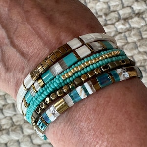 May include: A stack of bracelets with turquoise, white, gold, and brown beads. The bracelets are layered on a person's wrist.