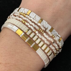 May include: A set of white and gold bracelets with different patterns and textures. The bracelets are stacked on a person's wrist.