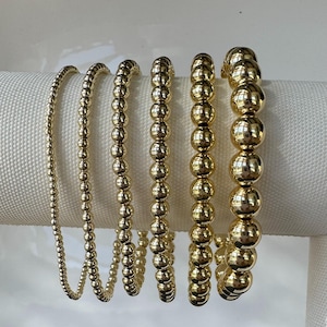 May include: Six gold beaded bracelets of varying sizes are displayed against a white background. The bracelets feature small to large, round beads, creating a layered look. The gold color is shiny and reflective, adding a touch of elegance.