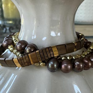 May include: Two stacked bracelets. The top bracelet features rectangular brown beads with gold accents. The bottom bracelet has large, dark brown, round beads. A third bracelet with gold-colored, geometric beads is also visible.