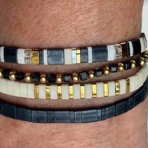 May include: A stack of four bracelets with black, white, and gold beads. The bracelets are layered on a person's wrist.