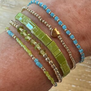 May include: A set of five stacked bracelets featuring a mix of light blue square beads, gold-toned round beads with a central heart charm, a wide lime green rectangular tile bracelet, a strand of gold-toned beads with green accents, and light blue beads.