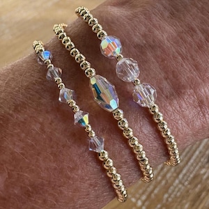 May include: Three stretch bracelets featuring small gold-toned metallic beads interspersed with clear, faceted crystal beads. The crystals exhibit an iridescent, rainbow-like sheen, and the bracelets are arranged in a stack on a wrist.