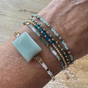 May include: A multi-strand bracelet set featuring a large rectangular light blue stone centerpiece. The design incorporates various small beads in shades of light blue, white, metallic gold, and dark gray, arranged in multiple rows on the wrist.