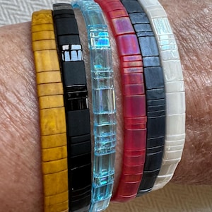 May include: A collection of six colorful bracelets. The bracelets are made of square, flat beads in various colors, including gold, black, iridescent blue, red, black, and white. The bracelets are worn on a wrist.