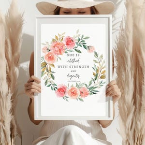 Può includere: Una stampa incorniciata bianca con una ghirlanda di fiori e il testo "Lei è vestita di forza e dignità. Proverbi 31:25"