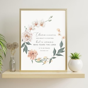 Op de afbeelding: Een ingelijste print met een gouden rand toont een bloemenkrans en scripttekst. De tekst luidt: "Charme is bedrieglijk, en schoonheid is vluchtig; maar een vrouw die de Heer vreest, is te prijzen. Spreuken 31:30."