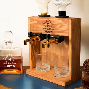 a wooden dispenser with two glass taps, a bottle of brown liquor, and two empty glasses on a wooden surface.