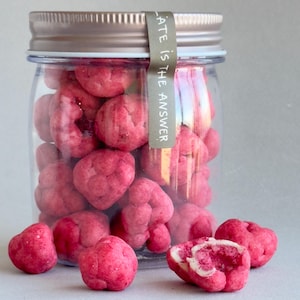 May include: A clear jar filled with heart-shaped, pink candies. The jar has a silver lid and a gray label that reads "Chocolate is the answer." Several candies are scattered around the jar.