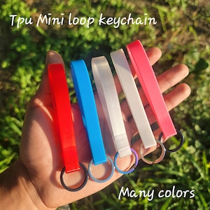 May include: A hand displays five TPU mini loop keychains in red, blue, white, and pink. Each keychain has a matching colored loop and a metal ring. The text "Tpu Mini loop keychain" and "Many colors" are visible.