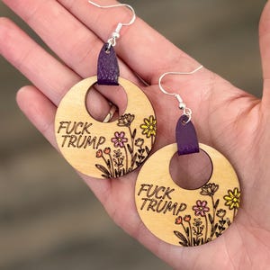 Op de afbeelding: Ronde houten oorbellen met de tekst "FUCK TRUMP" en bloemmotieven. Ze hebben paarse leren accenten en zilverkleurige haken. De oorbellen hebben een gat in het midden.