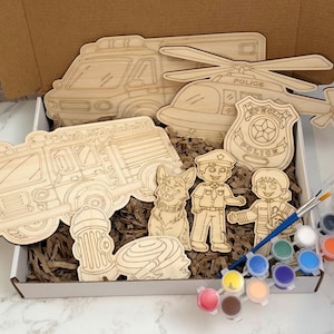 May include: A wooden craft kit featuring emergency service vehicles and characters. Includes a fire truck, ambulance, helicopter, police badge, fire hydrant, and a dog. Also includes paint pots and a paintbrush. The kit is packaged in a cardboard box.