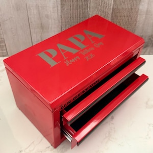 May include: A bright red metal tool chest with two drawers and a hinged lid. The top is engraved with the word PAPA and the text Happy Fathers Day 2026. The front of the drawer features the black text US GENERAL. The finish is glossy and smooth.