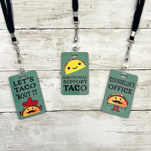 May include: Three green and white plastic badge holders with black lanyards. The badge holders feature a cartoon taco with a smiling face and text that reads "Let's Taco 'Bout It", "Emotional Support Taco", and "Counselor's Office".