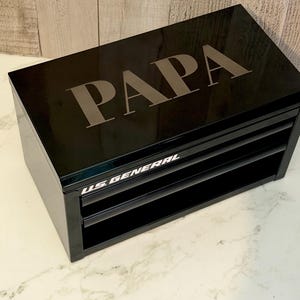 May include: A black metal tool chest with two drawers, featuring the word PAPA engraved in large capital letters on the top surface and the text U.S. GENERAL printed in white on the front edge of the top drawer frame.