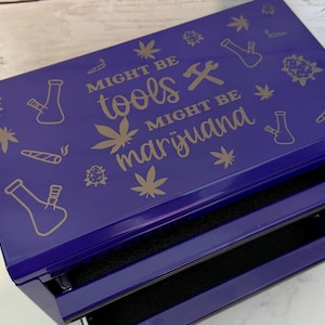 May include: A rectangular purple metal storage box with two pull-out drawers. The top surface features gold-colored line art of cannabis leaves, bongs, and joints, with the text "MIGHT BE tools MIGHT BE marijuana" printed in the center.