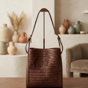 May include: A dark brown leather bucket bag featuring a woven exterior pattern. The bag has a structured rectangular base, a single adjustable shoulder strap attached with metal hardware, and a clean, open-top design with a consistent weave texture.
