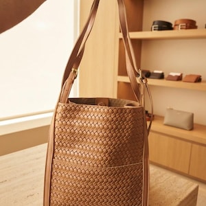 May include: A tan bucket bag featuring a woven exterior pattern, two long shoulder straps with adjustable metal buckles, and a structured rectangular base. The bag has a wide open top and a smooth, solid-colored trim along the vertical edges.