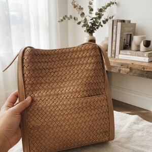 May include: A tan shoulder bag featuring a woven leather exterior with a rectangular silhouette. The bag includes a smooth leather border along the edges, a top opening, and a horizontal seam across the lower section creating a front pocket.