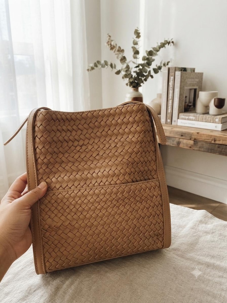 May include: A tan shoulder bag featuring a woven leather exterior with a rectangular silhouette. The bag includes a smooth leather border along the edges, a top opening, and a horizontal seam across the lower section creating a front pocket.