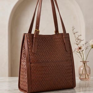 May include: A medium-sized brown tote bag featuring an intricate woven texture across the front and side panels. The bag includes two vertical shoulder straps with gold-toned metal buckles and a horizontal seam detail across the lower section.