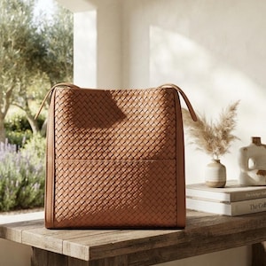 May include: A tan-colored tote bag featuring an intricate woven pattern across the front and back panels. The bag has a structured rectangular shape with smooth leather-like side panels and two thin shoulder straps attached at the top corners.