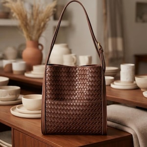 May include: A dark brown leather shoulder bag featuring an intricate woven pattern across the front panel. The bag has a vertical rectangular shape, a single long strap with a metal buckle adjustment, and smooth leather side panels with visible stitching.