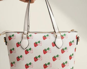 May include: A white handbag featuring a repeating pattern of red strawberries with green leaves. The bag has two white top handles, a gold-toned zipper closure along the top, and gold-toned hardware attachments for the handles on each side.