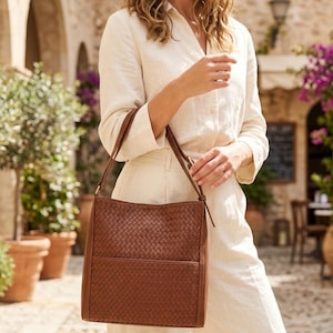 May include: A brown shoulder bag featuring a woven leather texture on the front panel and a smooth leather border. The bag has a rectangular shape, a single shoulder strap with a buckle adjustment, and a horizontal seam across the lower section.