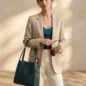 May include: A dark green woven leather tote bag with dual shoulder straps and a gold-tone side zipper, paired with a beige linen blazer, a dark green camisole, and cream-colored trousers. The bag features a structured rectangular shape.