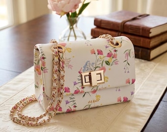 May include: White floral crossbody bag with gold chain strap and clasp. The bag features a pattern of pink, yellow, and purple flowers. The brand name "ALDO" is visible on the front of the bag.