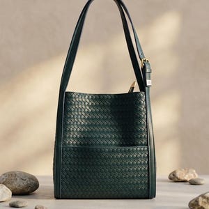 May include: A dark forest green bucket bag featuring an intricate woven leather exterior pattern. The bag includes a single adjustable shoulder strap with a gold-toned metal buckle and a top zipper closure with a matching leather pull tab.