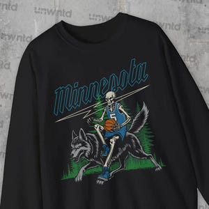Puede incluir: Sudadera negra con un gráfico de un esqueleto montando un lobo y sosteniendo una pelota de baloncesto. El texto "minnesota" está escrito en una fuente estilizada sobre el gráfico.