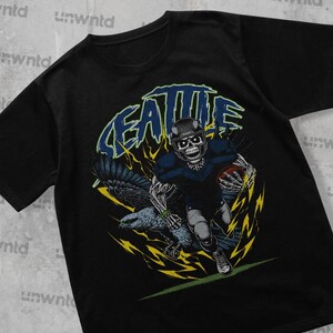 May include: Black t-shirt with a graphic of a skeleton football player wearing a blue jersey and helmet, running with a football. The graphic includes a black and white eagle with yellow lightning bolts in the background. The text "SEATTLE" is written in a stylized font above the player.