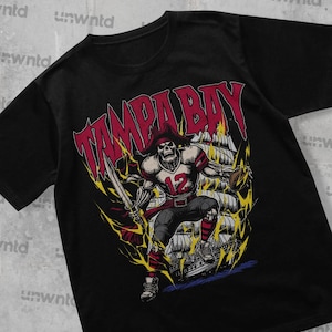 Puede incluir: Camiseta negra con un estampado gráfico de un esqueleto pirata que lleva una camiseta de fútbol con el número 12 y sostiene un balón de fútbol. El texto "TAMPA BAY" está impreso sobre el esqueleto en rojo y amarillo.