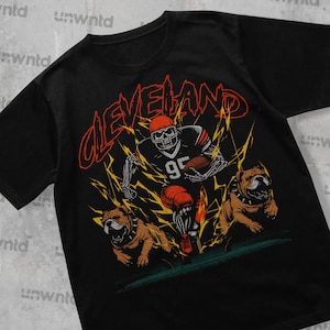 May include: Black t-shirt with a graphic print of a skeleton football player wearing a brown and orange jersey with the number 95. The player is running with a football and is surrounded by lightning bolts. Two bulldog dogs are running beside the player. The text "CLEVELAND" is printed in a bold, stylized font above the player.