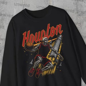 Może przedstawiać: Czarna bluza z nadrukiem przedstawiającym szkielet grający w koszykówkę przed rakietą z napisem "Houston" w czerwonych i żółtych literach.