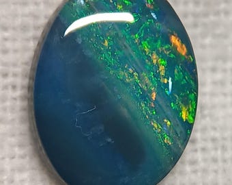 May include: Oval black opal gemstone with flashes of green, orange, and yellow. The polished surface reflects light, displaying the play-of-colour effect. The opal has a dark blue base colour.