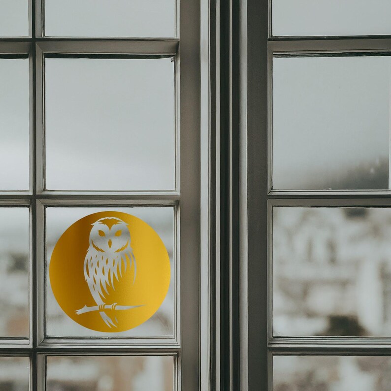 Op de afbeelding: Een raam met een gouden ronde sticker met een wit silhouet van een uil die op een tak zit.