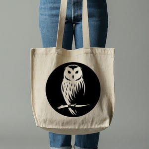 Op de afbeelding: Een beige tote bag met een zwart cirkelontwerp met een wit silhouet van een uil die op een tak zit.